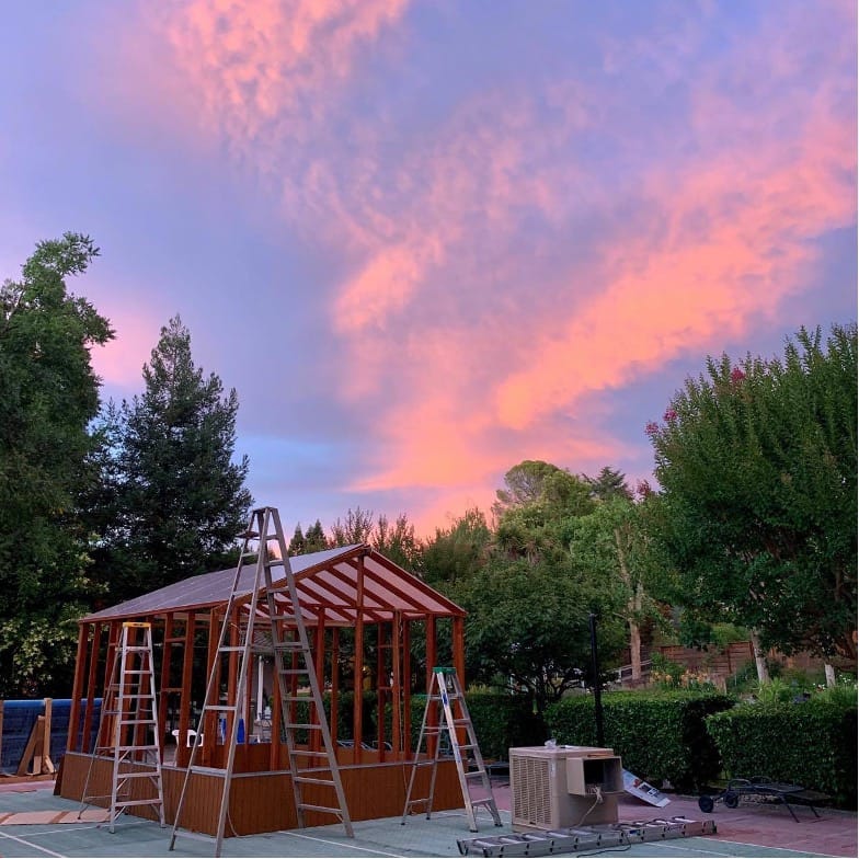 An under construction greenhouse, set next to trees underneath a pink and purple sky